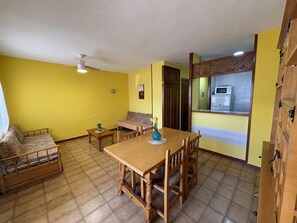 Un espace repas comprenant une table et des chaises en bois, un canapé et une cuisine visible à travers une cloison.