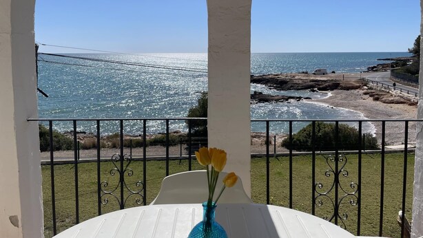 Ein Balkon mit Blick auf das Meer und eine Brücke. Ein weißer Tisch mit einer Vase voller gelber Blumen.