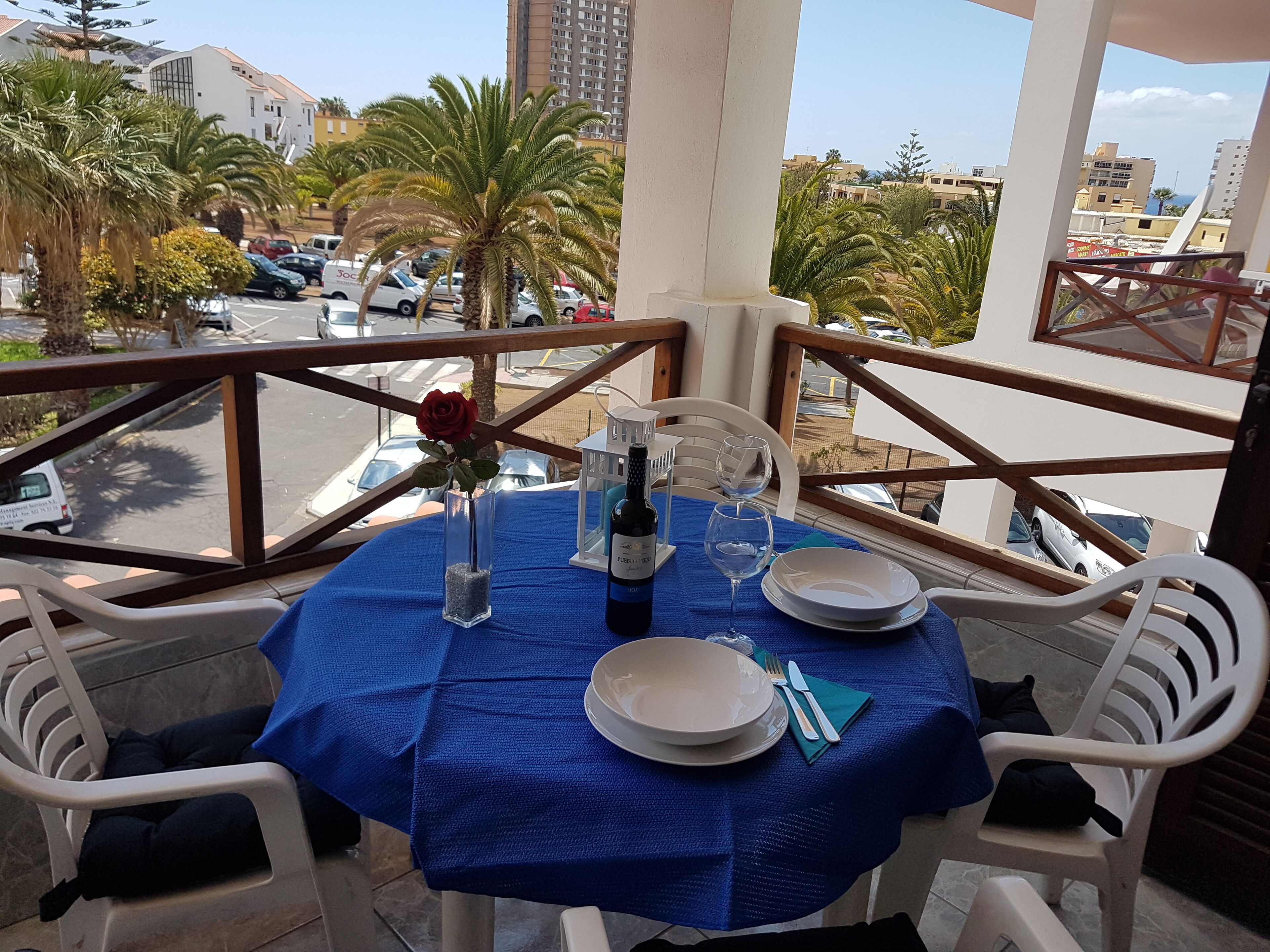 Une table de balcon dressée avec une nappe bleue, des assiettes blanches, une bouteille de vin et une rose rouge dans un vase en verre.