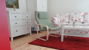Une pièce avec un plancher en bois, une commode blanche à tiroirs, une chaise verte, un canapé à motifs floraux et un tapis rouge.