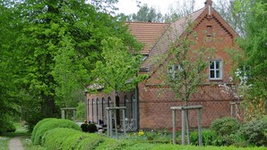 Une maison traditionnelle en briques, dotée d’un toit à pente, entourée d’une végétation luxuriante et bordée d’un sentier.