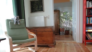 Un salon avec un fauteuil vert, une commode en bois, un tapis rouge, une bibliothèque et une plante en pot.
