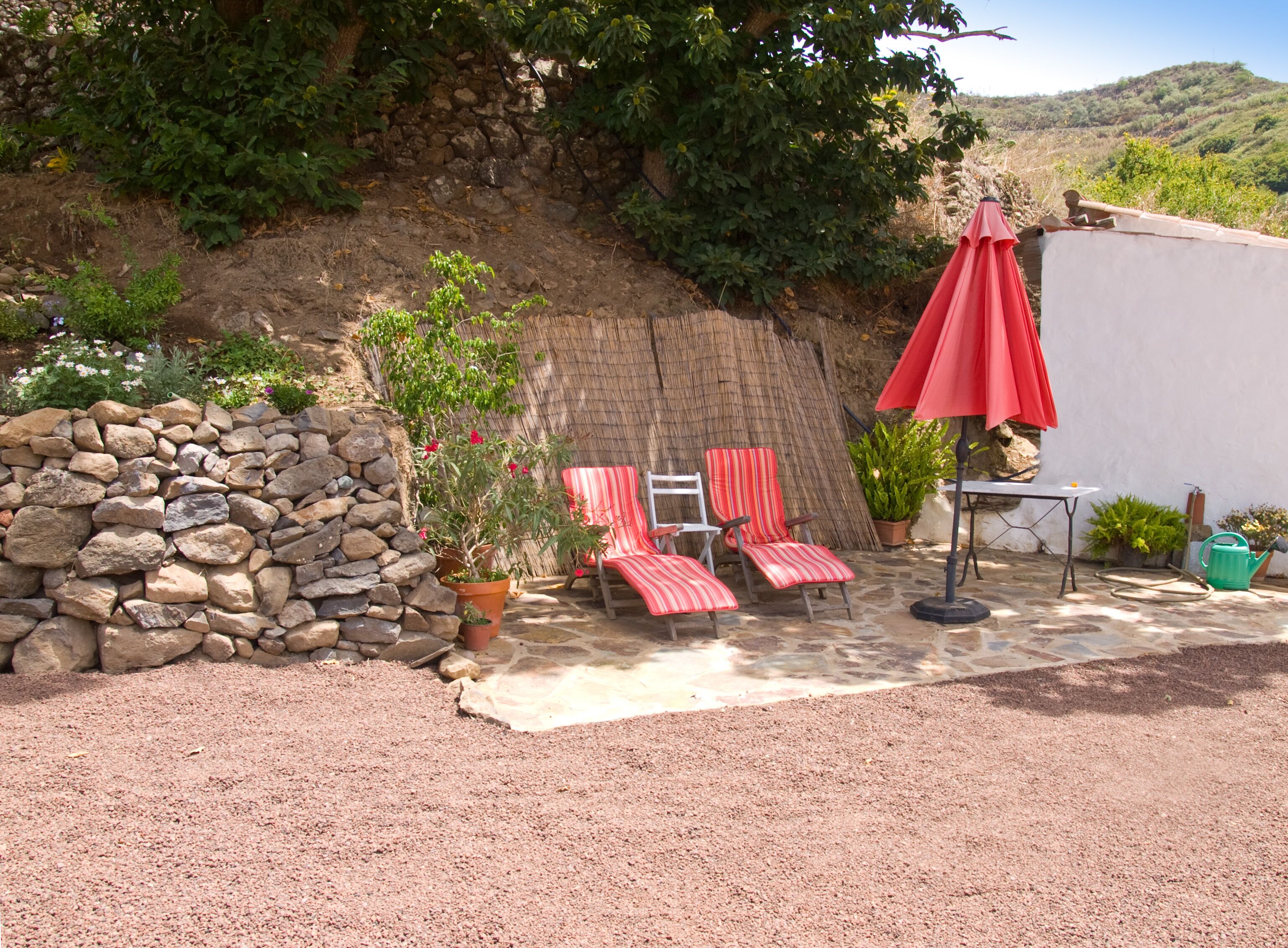 A patio with a stone wall, two striped lounge chairs, a red umbrella, and a table.
