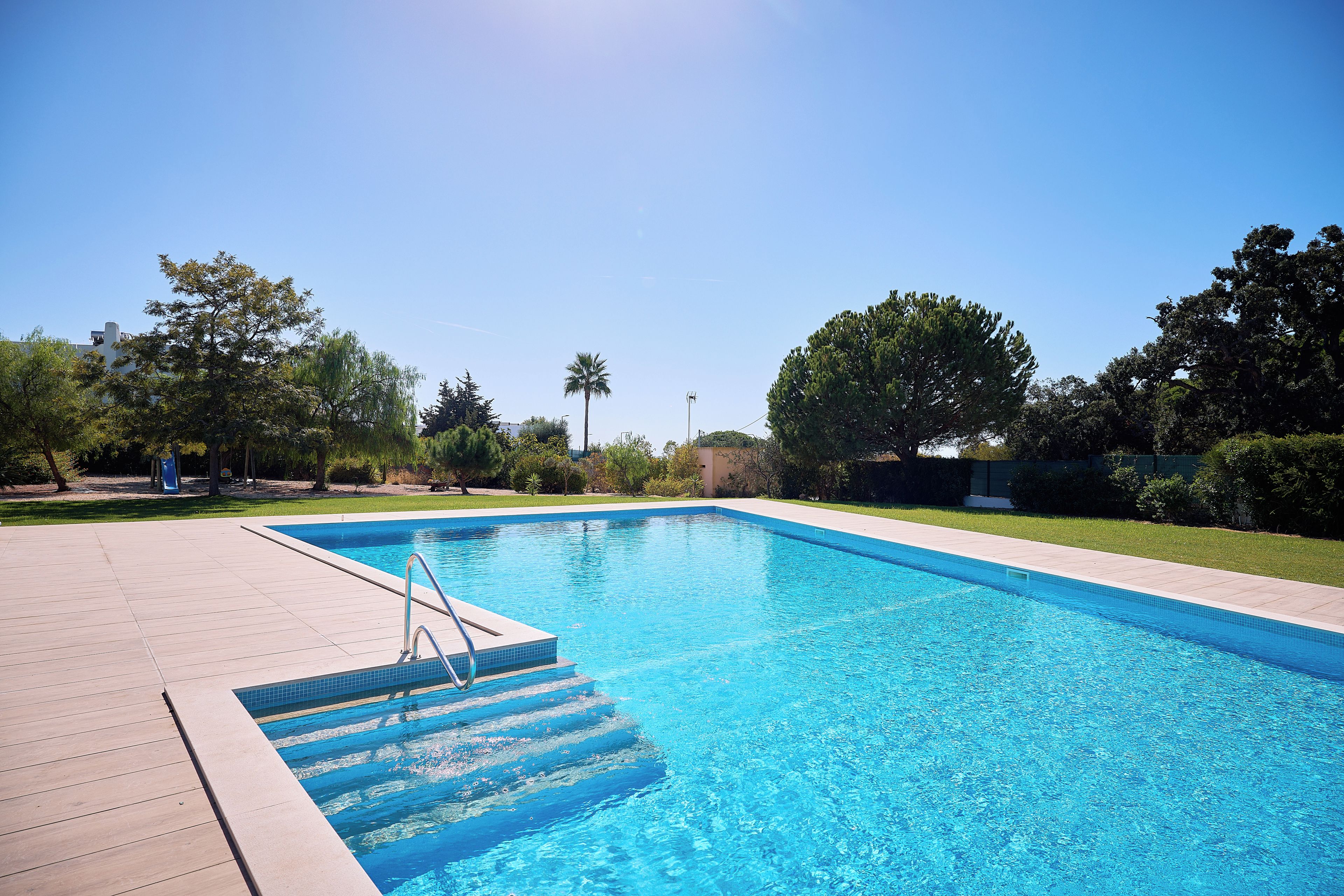 A rectangular swimming pool with clear blue water, surrounded by a tiled deck and a grassy area with trees.