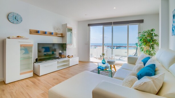 Una sala de estar moderna con televisor, muebles blancos, un reloj y un balcón con vista al mar.
