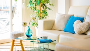Una sala moderna con un sofá, una mesita de centro de vidrio, un taburete pequeño, una planta en maceta y un jarrón con flores.