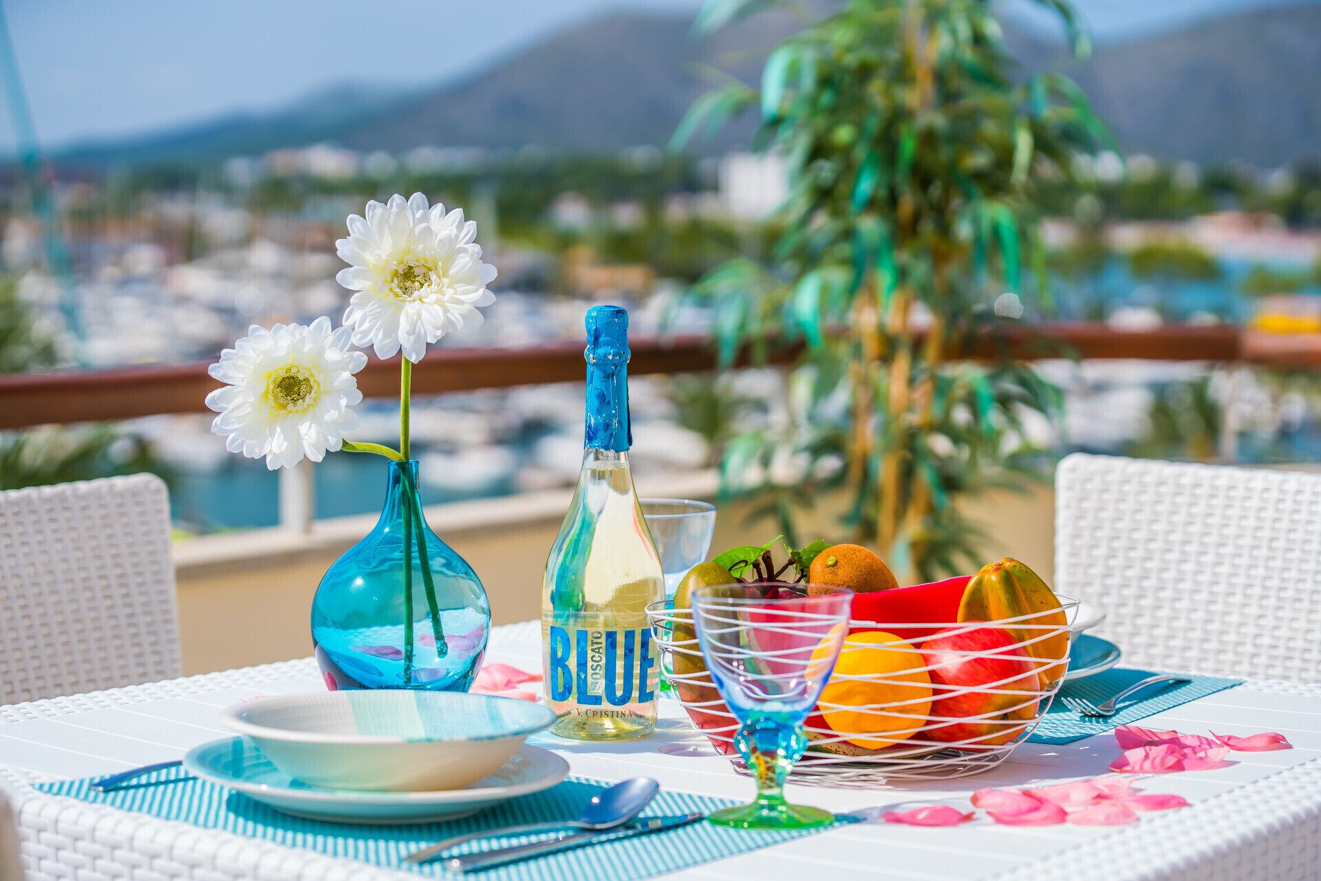 Une table avec une bouteille de Bia, un verre d’eau, un bol et un panier de fruits, le tout placé sur un balcon donnant sur une marina.