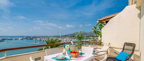 A balcony with wicker furniture, a table set with a bowl of fruit, and a view of a marina with boats and a mountain in the background.