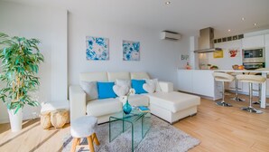Una sala de estar moderna con un sofá blanco, cojines azules, una mesa de centro de vidrio y una planta grande.