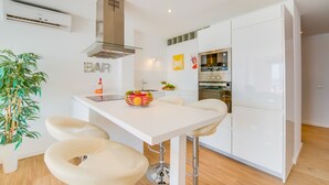 Una cocina moderna con armarios blancos, horno empotrado y refrigerador con puerta de vidrio. Cuenta con una zona de bar con taburetes y un cuenco de fruta sobre la encimera.