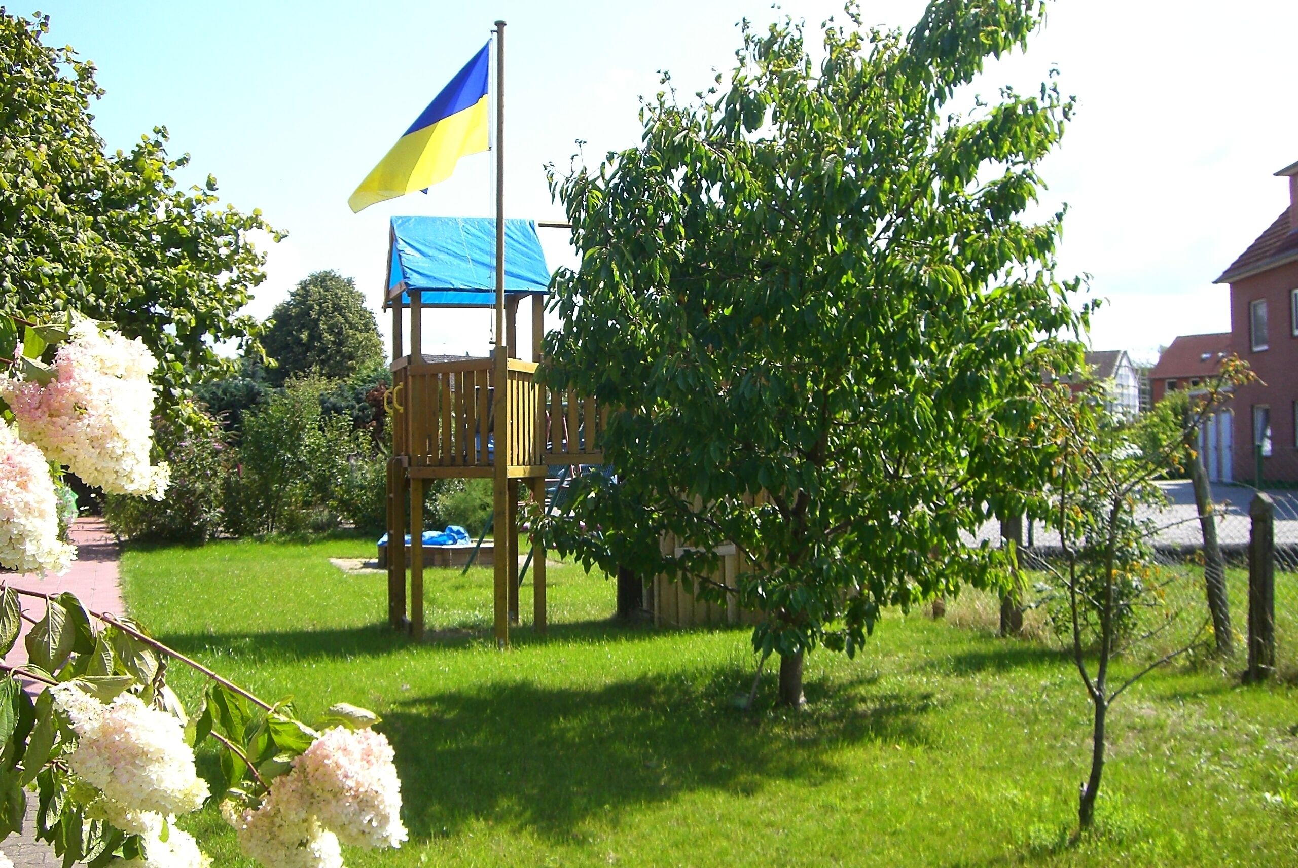 A wooden play structure with a blue tarp roof, a flagpole with a blue and yellow flag, and a tree in the foreground.
