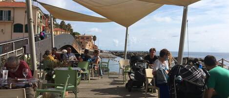Un área de comedor al aire libre con mesas y sillas, vista al mar y edificios al fondo.