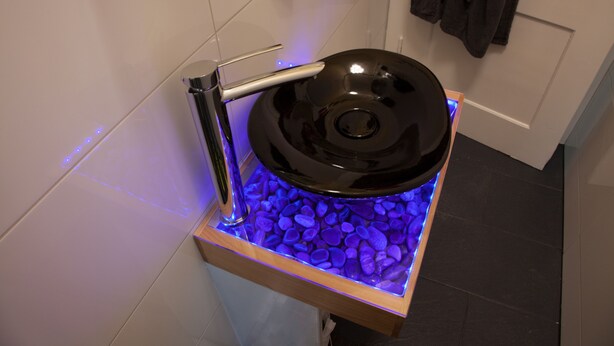 Ein modernes Badezimmer mit einem einzigartigen Spülendesign, bestehend aus einer schwarzen Spüle auf einem Holzfuß, umgeben von von blauen Lichtern belebten Kieselsteinen.