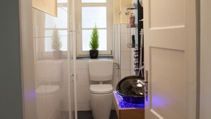 Ein schmales Badezimmer mit Toilette, einem Waschbecken mit blauer Beleuchtung, einer Topfpflanze und einem Fenster.