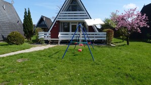A red and white chalet with a triangular roof, a white porch, and a swing set in the yard. - The family-friendly Villa Linus is located in a vacation park behind the dyke (Butjadingen)