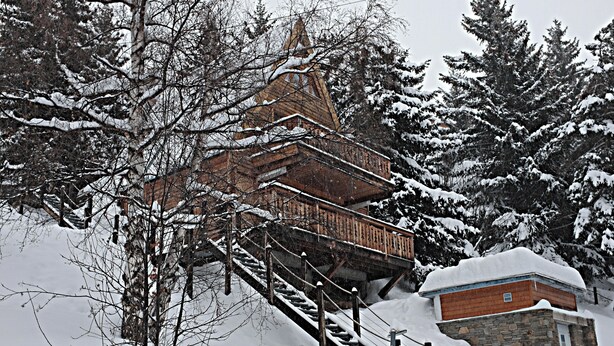 Una cabaña de madera con una escalera pronunciada, rodeada de árboles cubiertos de nieve y un edificio de piedra.