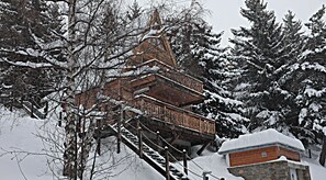 A wooden chalet with a steep staircase, surrounded by snow-covered trees and a stone building.