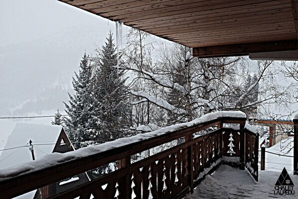 A wooden balcony with snow-covered railings overlooking a snowy landscape with trees and a building in the background.
