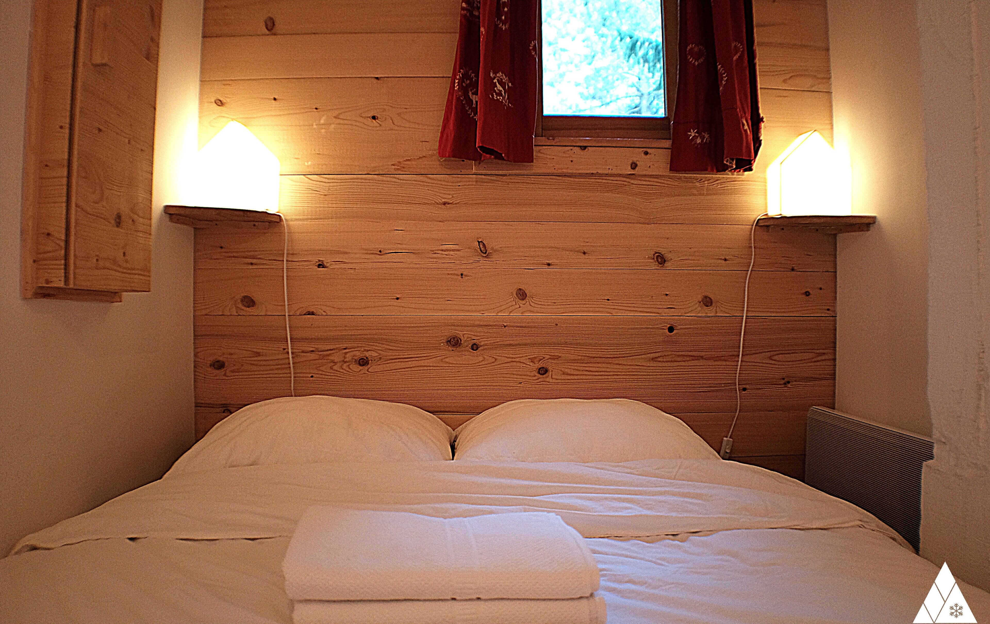 A wooden headboard with two wall-mounted lamps, a window with red curtains, and a neatly made bed with pillows and a folded blanket.