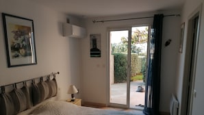 Een slaapkamer met een bed, nachtkastje, lamp, fotolijst, airconditioning en een schuifdeur naar een tuin.