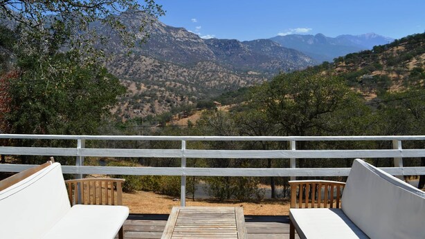 Une terrasse dotée de deux bancs blancs rembourrés et d’une table en bois, offrant une vue sur un paysage montagneux parsemé d’arbres et sous un ciel dégagé.