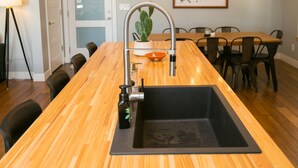 A modern kitchen with a wooden countertop, a sink, and a dining area with chairs and a table. - Downtown Mountain Views Craftsman Stunner 5 bd/4b (Fort Collins)