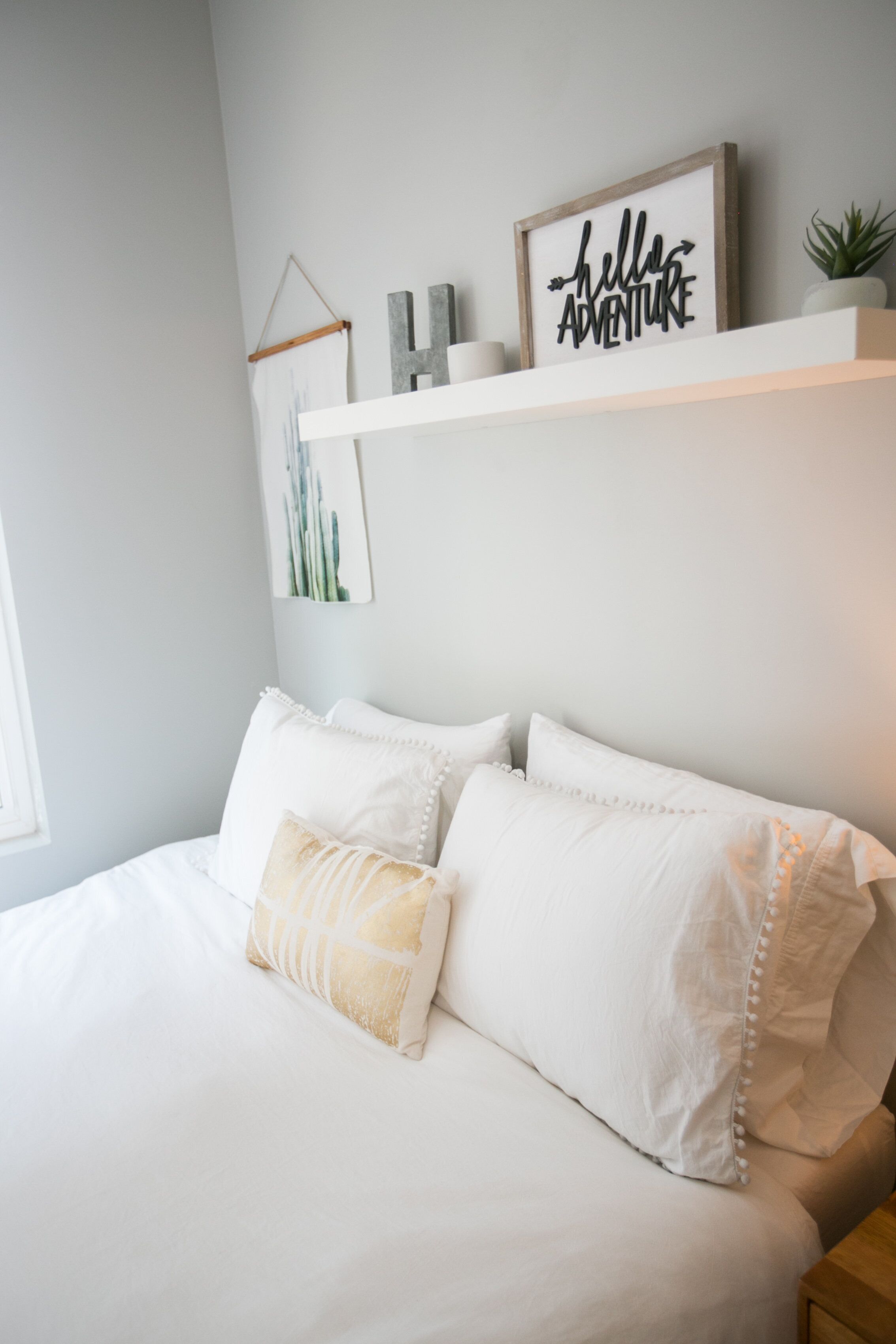 A neatly made bed with white bedding and decorative pillows, a small plant on a shelf above, and a framed sign that reads "hello ADVENTURE" on the wall.