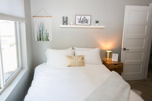 A neatly made bed with white bedding and decorative pillows, a bedside table with a lamp, and a wall-mounted shelf with a framed sign and a cactus painting. - Downtown Mountain Views Craftsman Stunner 5 bd/4b (Fort Collins)