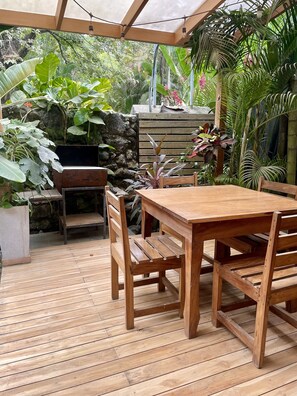 A wooden deck with a table and chairs, surrounded by lush tropical plants and a stone wall. - Spacious Villa Pool/Patio/Garden min from the Beach. H speed internet (Playa Santa Teresa)