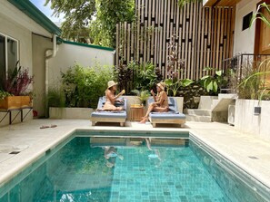 A swimming pool with two lounge chairs and a small table, surrounded by greenery and a wooden fence. - Spacious Villa Pool/Patio/Garden min from the Beach. H speed internet (Playa Santa Teresa)