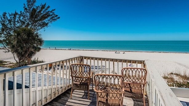 A beach view with a wooden deck, wicker chairs, and a palm tree.