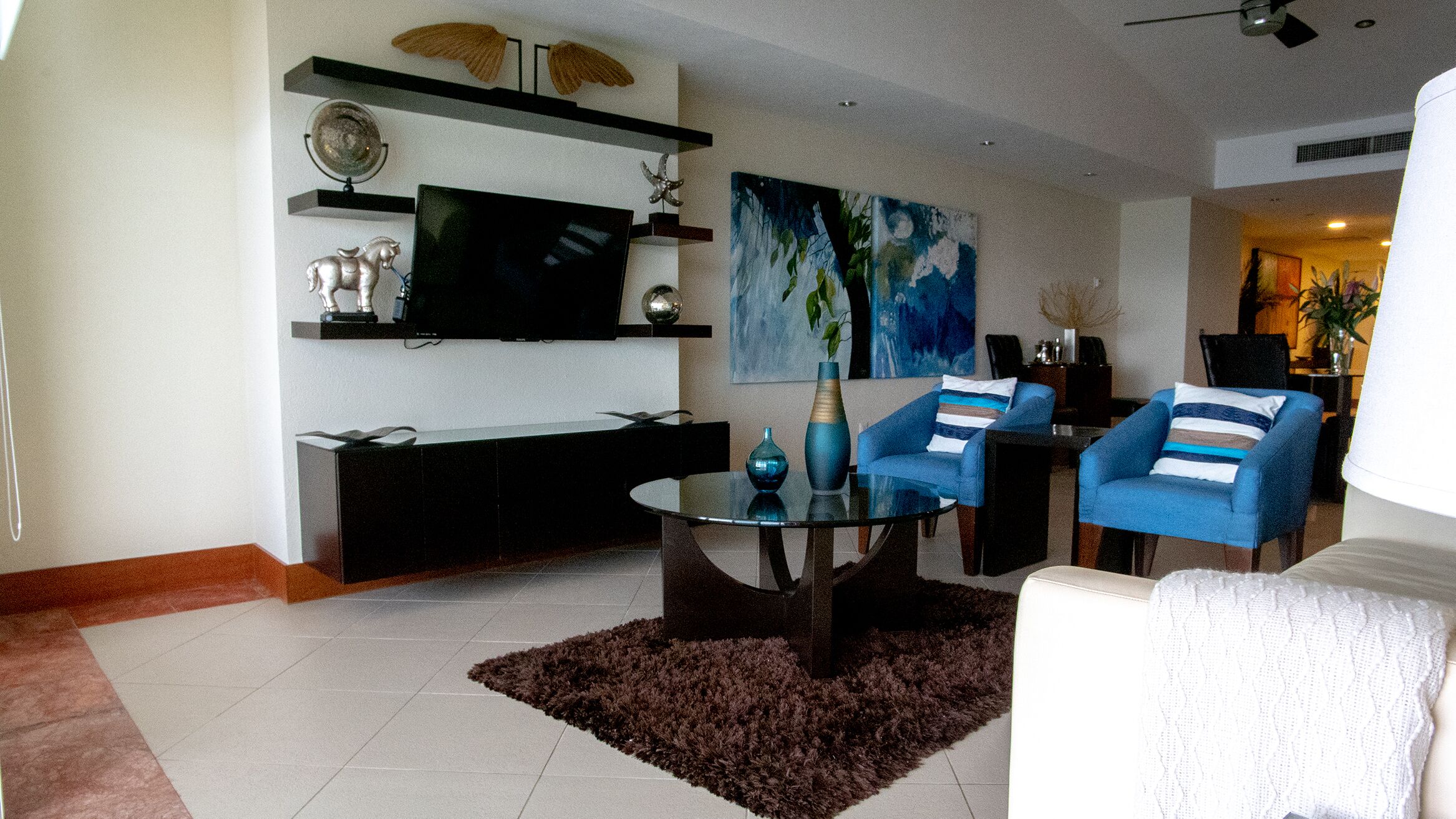 Un salon moderne avec un téléviseur à écran plat, des fauteuils bleus, une table basse et des objets décoratifs disposés sur des étagères.