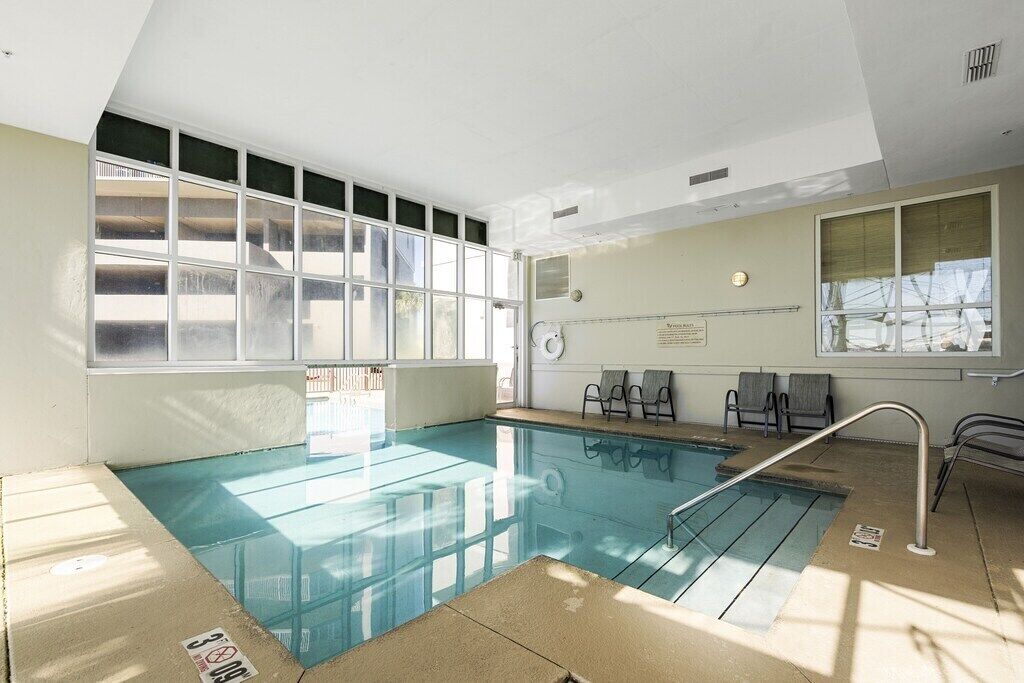 An indoor swimming pool with clear glass walls, a tiled floor, and a few lounge chairs.
