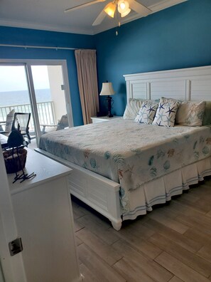 A bedroom with a bed, a nightstand, a lamp, a chair, and a view of the ocean.