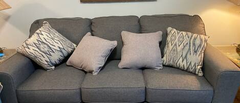 Un canapé gris avec trois coussins à motifs, un tableau encadré au-dessus, et une lampe sur la droite.