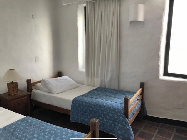 A small, simple room with two single beds, a nightstand with a lamp, a window with curtains, and a wall-mounted light fixture.