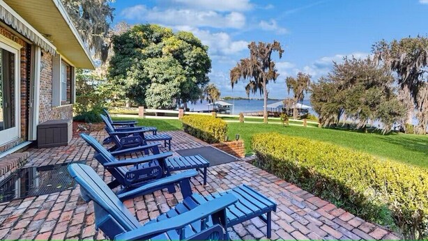 A patio with blue chairs and a brick floor, surrounded by greenery and trees. - Large beautiful house on lake. Dock, Water trampoline, Fire Pit, Rope Swing, 4/3 (Lake Placid)