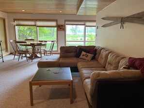 A cozy living room with a sectional sofa, a wooden coffee table, and a dining area with a table and chairs. Large windows provide a view of the outdoors.