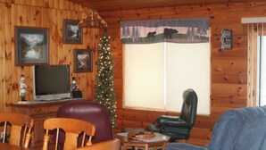 A cozy cabin interior with wooden walls, a dining area with wooden chairs, a leather sofa, a Christmas tree, and a television on a wooden stand. - Bears Den on the private lake (Winter)