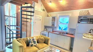 A cozy living room with a sofa, a glass coffee table, and a patterned rug. There is a kitchen area with a microwave and a refrigerator. A spiral staircase is visible in the background. - Carriage House, Forsyth Park, Pet friendly, private courtyard 🩷 (Savannah)