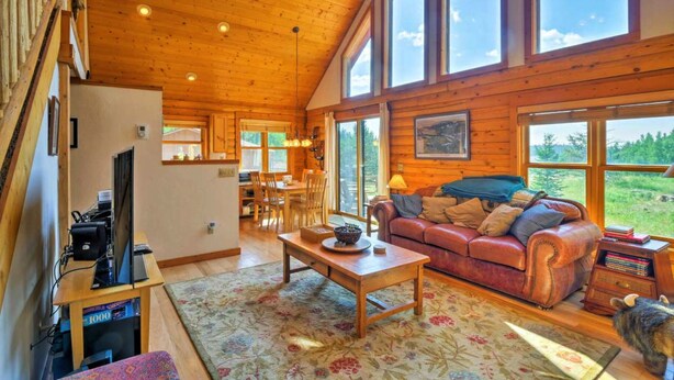 Interior acogedor de una cabaña con techo de madera, una sala de estar con sofá y mesa de centro, un comedor y grandes ventanales con vistas al exterior.