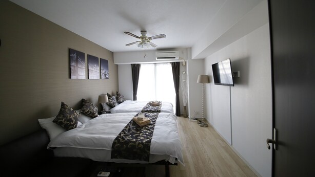 A hotel room with two beds, a ceiling fan, an air conditioning unit, a flat-screen TV, and a door. - Best Residence Kujo 502 (Osaka)