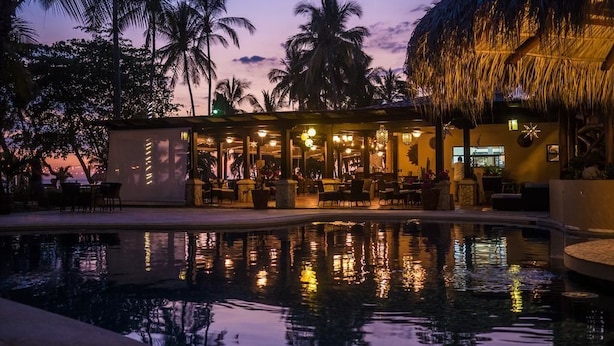 A poolside area with a bar and seating, surrounded by palm trees and a thatched-roof structure. - Sunset del Mar Beach Front House (Parrita)