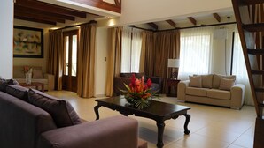 A living room with a brown sofa, a beige sofa, a coffee table with a floral centerpiece, and a lamp on a side table. - Sunset del Mar Beach Front House (Parrita)