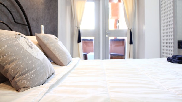 A neatly made bed with a white comforter and two decorative pillows. A window with curtains is visible in the background, and an air conditioning unit is mounted on the wall. - El Hamra Hotel (Marrakech)