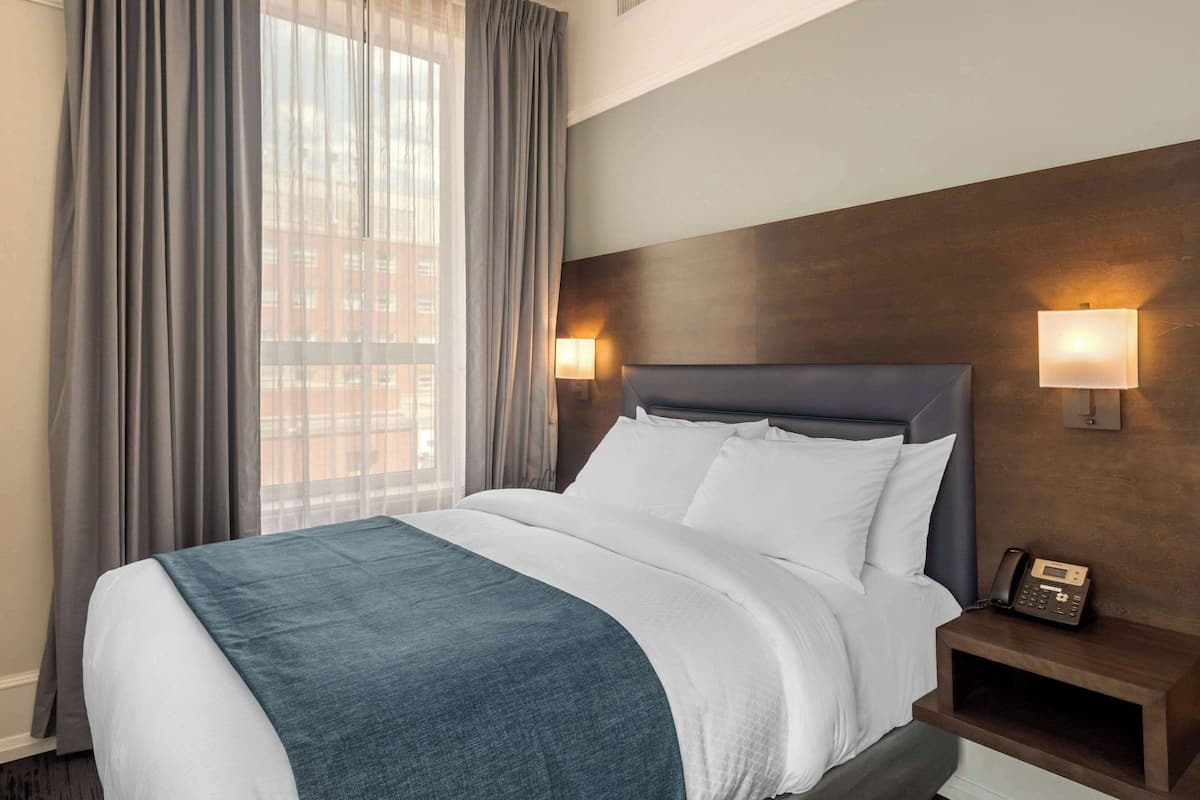 A hotel room with a large bed, grey curtains, a wooden headboard, two bedside lamps, and a telephone on the nightstand.
