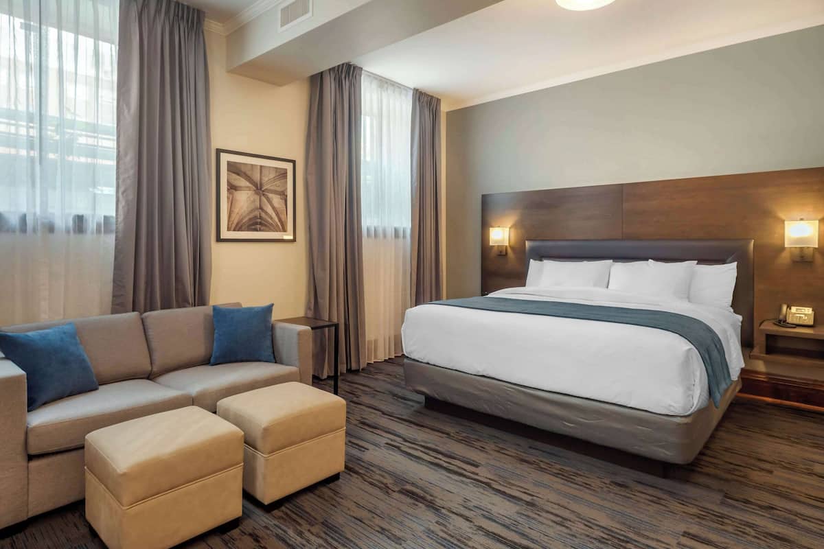 A hotel room with a large bed, a sofa, two ottomans, a small table, and a nightstand with a lamp.
