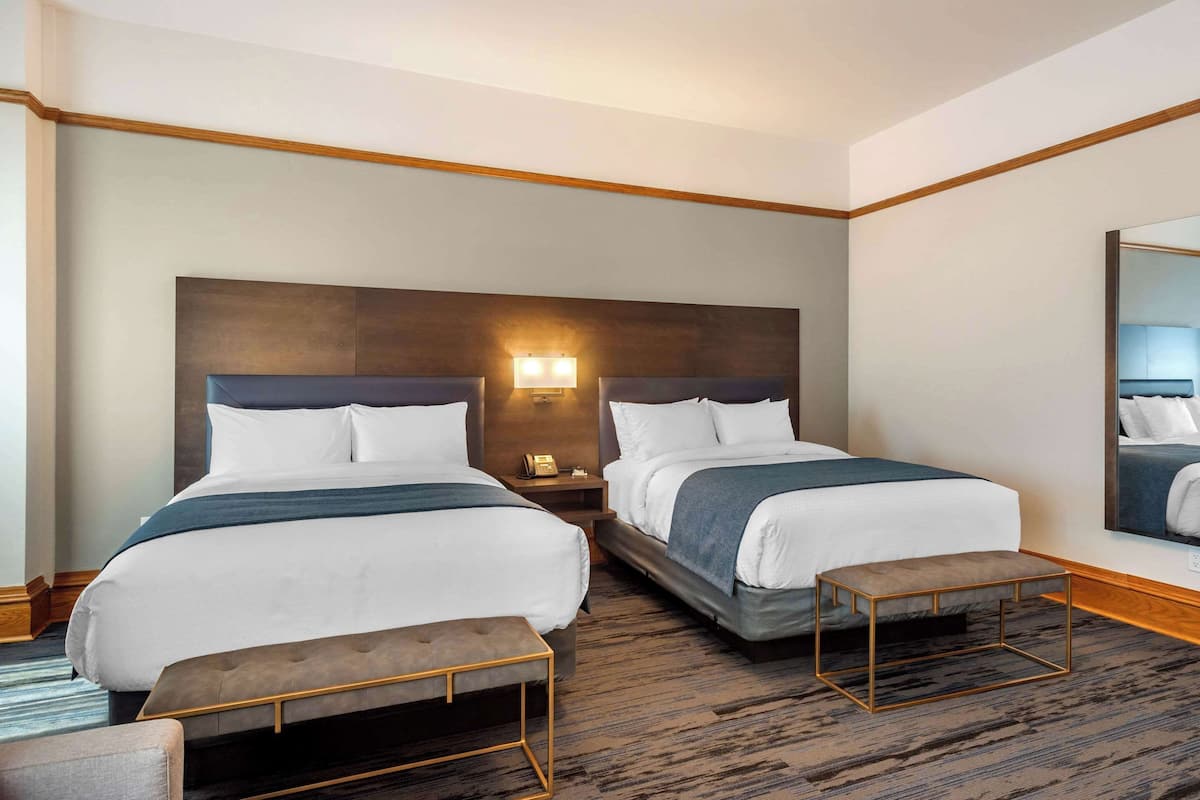 A hotel room with two beds, a nightstand with a lamp, a bench, and a large mirror on the wall.