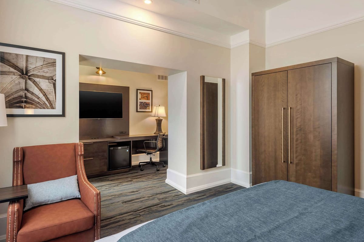A hotel room with a bed, a brown leather chair, a desk with a lamp, a TV, and a wardrobe.