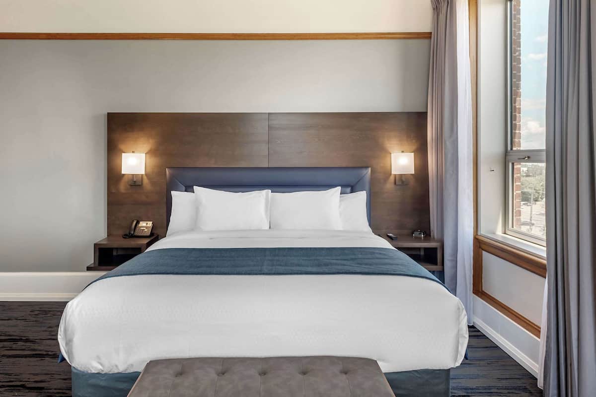 A hotel room with a large bed, two bedside tables with lamps, a wooden headboard, and a window with curtains.
